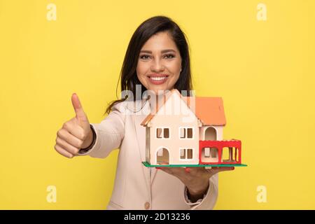 young woman in pink suit making thumbs up gesture and showing building model smiling on yellow background Stock Photo