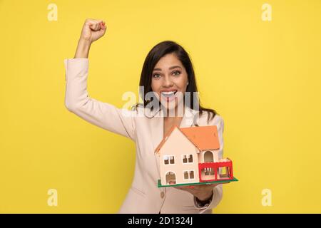 excited young girl in pink suit holding fist up and presenting house model, laughing and cheering victory on yellow background Stock Photo