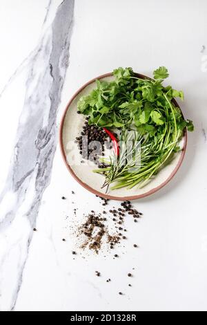 Black pepper peppercorns, ground powder, chili pepper and fresh herbs in ceramic plate over white marble background. Top view, space. Stock Photo