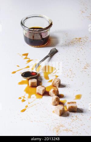 Homemade liquid transparent brown sugar caramel in glass jar with spoon and cane sugar cubes over white marble background. Copy space. Stock Photo