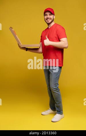 Happy man from delivery service in red t-shirt and cap giving food order and holding pizza box isolated over yellow Stock Photo