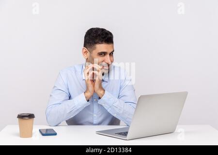 Sneaky scheming businessman sitting office workplace with laptop on desk, clasping hands and smirking mysteriously, having tricky cunning plan in mind Stock Photo