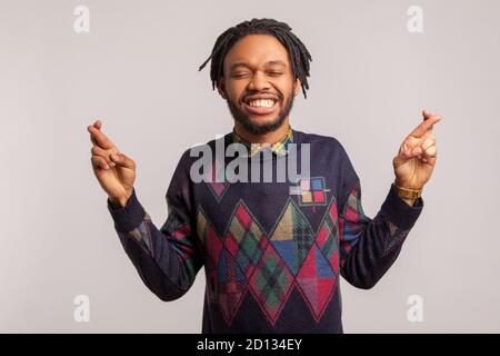 Positive african man with beard and dreadlocks crossing fingers and closing eyes making wish with toothy smile on face, rituals. Indoor studio shot is Stock Photo