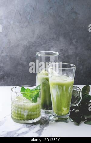 Matcha green tea iced latte or cocktail in three different glasses with ice cubes, matcha powder and jug of milk on white marble table, decorated by g Stock Photo
