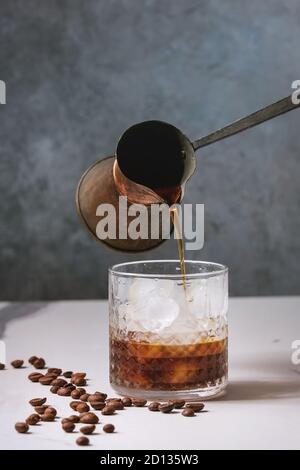 Iced coffee cocktail or frappe with ice cubes and cream served in glass with pouring coffee from vintage jezva and coffee beans around on white marble Stock Photo