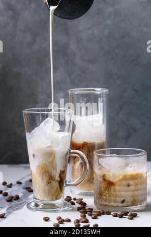 Iced coffee cocktail or frappe with ice cubes and pouring cream served in three different glasses with coffee beans around on white marble table with Stock Photo