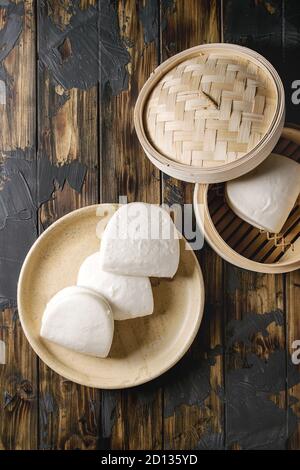 Empty gua bao steamed buns in ceramic plate and opened bamboo steamer over dark wooden plank background. Flat lay, space. Asian fast food. Stock Photo