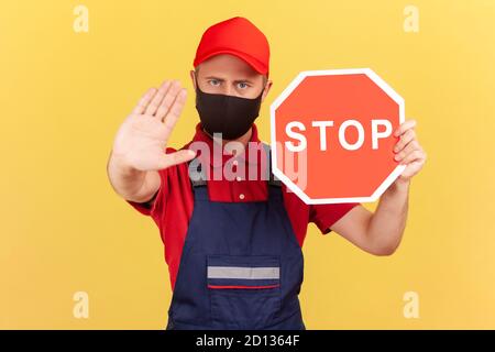 Craftsman in uniform and hygienic face mask showing stop gesture and holding red stop road sign, prohibitions and restrictions during quarantine. Indo Stock Photo