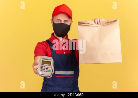 Courier in uniform and black protective mask showing paper bag and holding payment terminal, fast delivery and contactless payments on quarantine. Ind Stock Photo