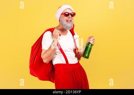 Happy elderly man in santa claus hat, trendy sunglasses and pants with suspenders holding bottle with alcohol and big red bag, celebration, bad habit. Stock Photo