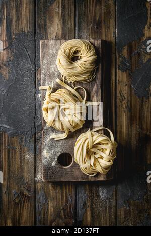 Variety of italian homemade raw uncooked pasta spaghetti and tagliatelle with semolina flour on wood cutting board over dark plank texture wooden tabl Stock Photo