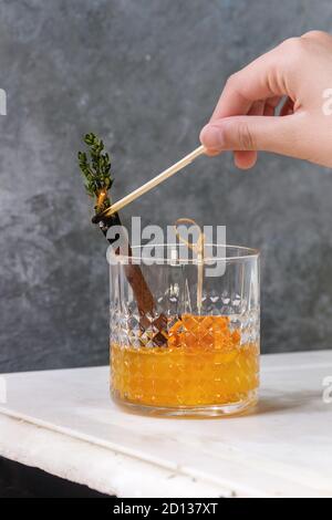 Glass of Scotch Whiskey orange juice alcohol cocktail with swirled orange peel on skewer, thyme and cinnamon sticks set fire to match, on white marble Stock Photo