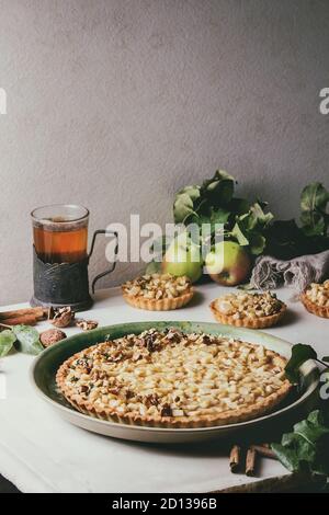 Homemade sweet apple shortbread tart in green ceramic plate, mini tartlets and hot tea in vintage cup holder with cinnamon sticks, walnuts, apples bra Stock Photo