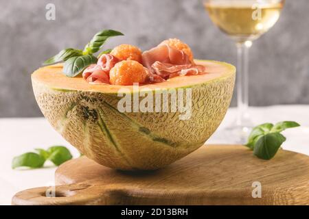 Melon and ham or prosciutto salad served in half of Cantaloupe melon, decorated by fresh basil standing on wooden serving board over white tablecloth Stock Photo