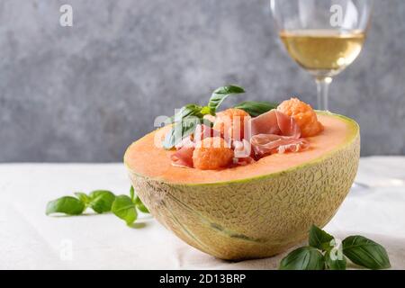 Melon and ham or prosciutto salad served in half of Cantaloupe melon, decorated by fresh basil standing on white tablecloth with glass of white wine. Stock Photo
