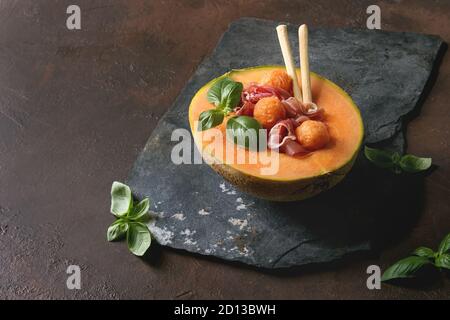 Melon and ham salad served in half of Cantaloupe melon with fresh basil and grissini bread on black slate board over dark brown texture background. Stock Photo