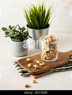 A lot of cashew nuts in a glass container on a wooden Board on a light background. green plants in pots. Stock Photo