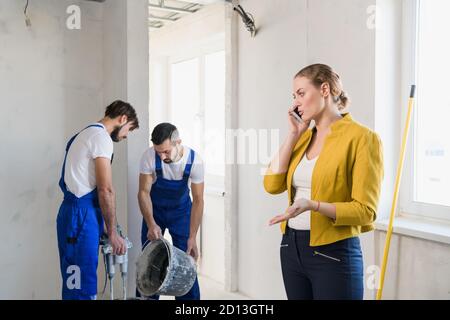 Two construction workers are preparing cement in a bucket. Woman speaks seriously on the phone Stock Photo