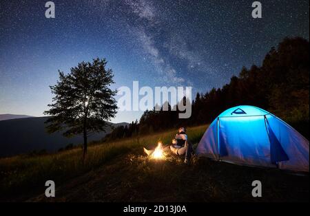 Young romantic couple travellers resting at campfire near illuminated blue tourist tent under amazing night sky full of stars and Milky way. On background beautiful starry sky, mountains and big tree Stock Photo