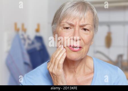 Acute toothache: senior woman in her kitchen with a hand at her painful jaw. Stock Photo