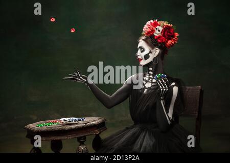 Gambling. Young girl like Santa Muerte Saint death or Sugar skull with bright make-up. Portrait isolated on dark green studio background with copyspace. Celebrating Halloween or Day of the dead. Stock Photo
