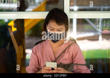 Asian woman wearing protective mask on her face while being and using smartphone in the coffee shop during virus COVID-19 pandemic. Stock Photo