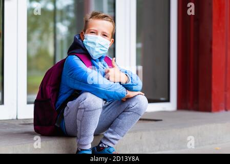 Cute looking at camera school boy 9s old sit on the stairs wears casual clothes and backpack face mask near school. Kid shows thumbs up Stock Photo