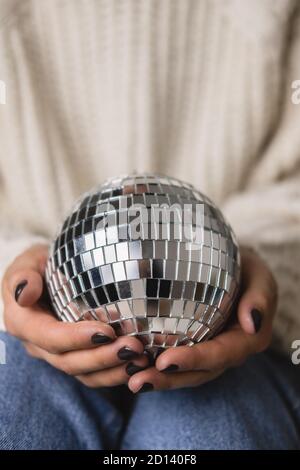 Young woman is sitting on the bed in cozy knitted woolen white sweater, socks and holding toy disco ball in her hands. Hygge, New Years, Christmas, ho Stock Photo