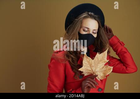 Life during covid-19 pandemic. middle aged woman in red coat with black mask and yellow autumn maple leaf isolated on beige. Stock Photo