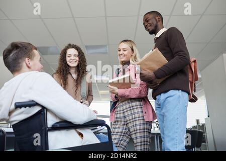 Low angle view at multi-ethnic group of students talking to young man in wheelchair while studying together in college library