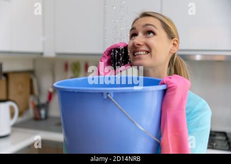 apartment flooded by upstairs - worried woman calling repair service while water leaking from ceiling in bucket Stock Photo