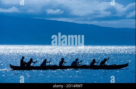 A six-person team of paddlers practicing for a race in a traditional Hawaiian outrigger canoe is silhouetted by sunlight glistening on the North Pacific Ocean off the coast of Maui in Hawaii, USA. Typical Hawaiian racing canoes have a single outrigger on one side of the craft that provides stability to the long sleek boat. Three crew members pull through the water on each side with paddles that feature long handles with a single blade at one end. Also seen here is a seventh man, a coach who sits higher in the stern (rear) of the boat during practice but is never aboard during actual races. Stock Photo
