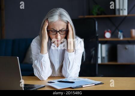 Burnout and overwork concept.Tired businesswoman working overtime in office.Portrait of stressed grey-haired senior woman with head ache.Eye strain Stock Photo