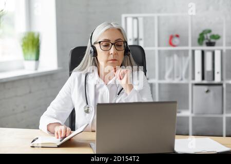 Portrait of senior grey-haired female doctor in her office using laptop for video chat with a patient. Online consultation with doctor for diagnoses Stock Photo