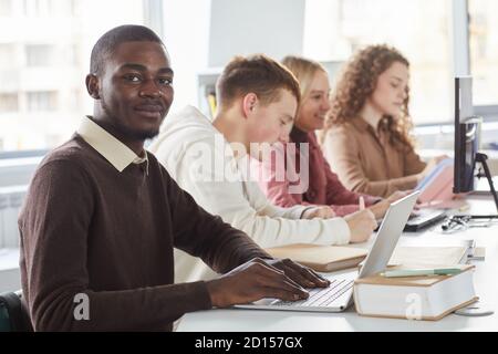 Portrait of smiling African-American man using laptop and looking at camera while studying with group of students in college Stock Photo
