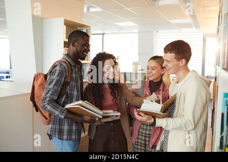 Waist up portrait of multi-ethnic group of students chatting cheerfully while standing by shelves in school library lit by sunlight Stock Photo