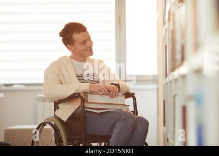 Warm toned portrait of young man using wheelchair in school while looking at bookshelves in library lit by sunlight Stock Photo