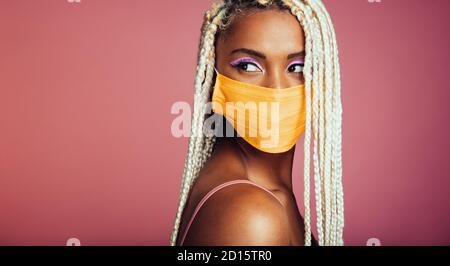 Young african woman with blonde box braids wearing protective face mask. Female with african hair style and face mask looking away on pink background.