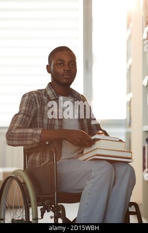 Vertical warm toned portrait of young African-American man using wheelchair in school while looking at camera in library lit by sunlight Stock Photo