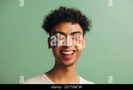 Close-up of a cheerful young man with pride flag painted on face. Gay man with rainbow face paint smiling against pastel green background. Stock Photo