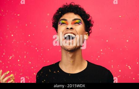 Cheerful gay man with glitters flying against red background. Excited young male with rainbow eye makeup throwing up the glitter and laughing. Stock Photo