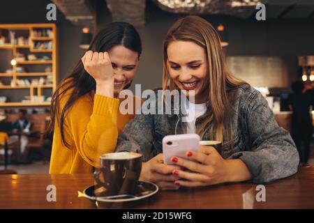 Happy female friends at a cafe using smart phone with a cup of coffee on the table. Women friends looking at a mobile phone and laughing. Stock Photo