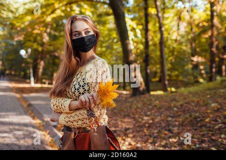Pretty young woman wears protective mask walking in autumn park holding leaves. Coronavirus covid-19 fashion Stock Photo