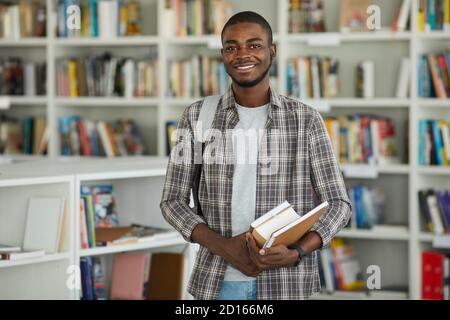 Waist up portrait of young African-American man in school library holding books and smiling happily at camera, copy space Stock Photo