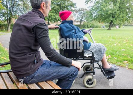 Carer (son) with 90 year old Mother in wheelchair in public park. UK Stock Photo