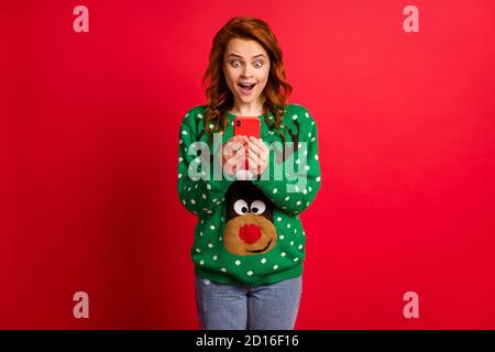 Photo portrait of excited girl with open mouth holding phone in two hands wearing ugly green sweater isolated on vivid red colored background Stock Photo