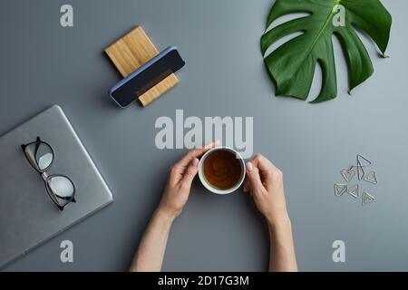 Above view flat lay of female hands holding coffee cup over grey workplace background with business gadgets and tropical leaf, copy space Stock Photo