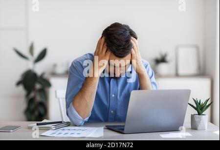 Business Problems. Desperate Millennial Businessman Sitting At Workplace In Office, Touching Head Stock Photo