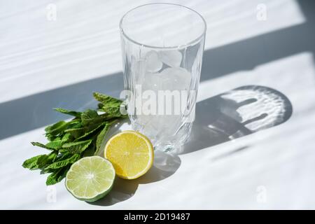 Ingredients for making mojito on white background. lime, lemon, mint leaves and high glass with ice cubes. space for text. trendy direct sun light. summer non-alcoholic cocktail. Stock Photo