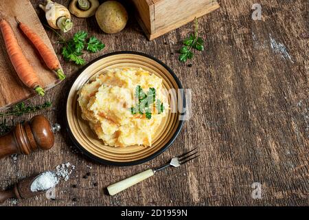 Delicious and healthy mashed parsnips with potatoes and carrots and ingredients for making vegetarian parsnip puree on a dark wooden background. Top v Stock Photo